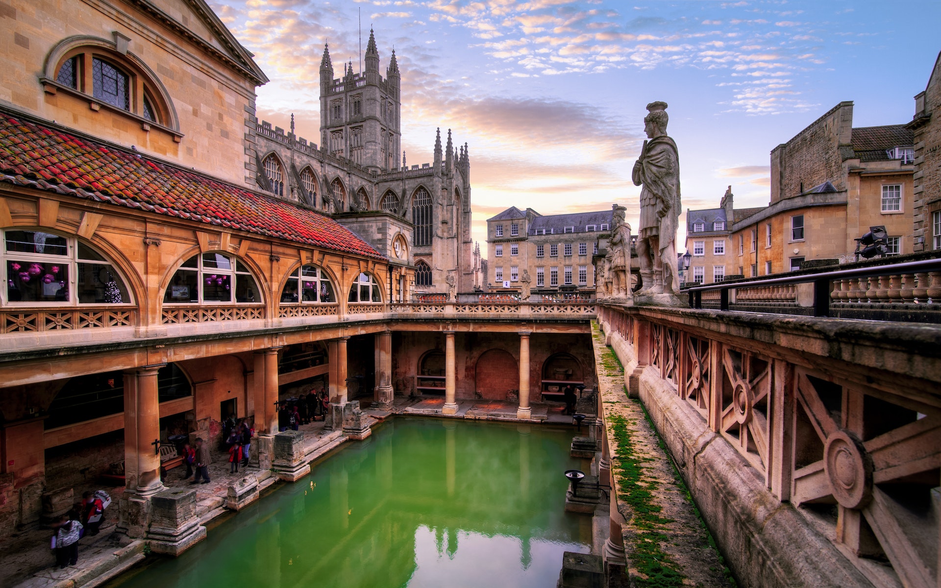 People tour a green rectangle of water surrounded by a Roman structure with a large abbey in the background